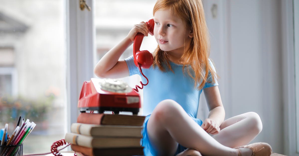A Question about the little girl in red dress in Schindler's List - Low angle of calm redhead preteen lady in blue dress and beige sandals looking away and having phone call using retro disk telephone on stack of books while sitting with legs crossed on wooden table against window at home