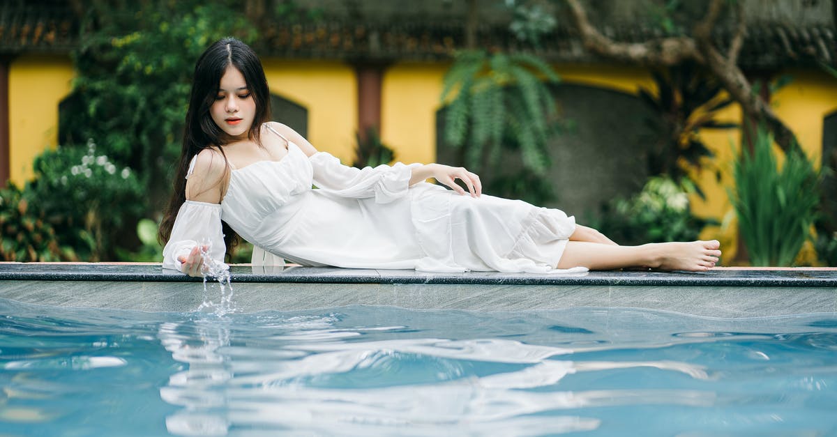 After the end of Edge of Tomorrow, would Cage be caught in a time-loop? - Full body of thoughtful barefoot Asian female in white dress looking down while lying on poolside near clear water near building and tree