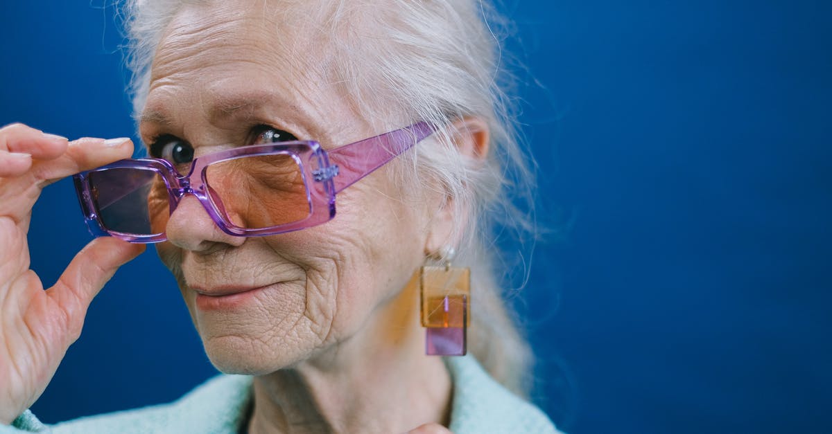 Age issue in How Old Are You - Portrait of elegant smiling gray haired elderly female wearing purple sunglasses and earrings looking at camera against blue background
