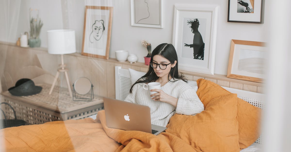 Alternate Versions of Movies - Focused young brunette in eyeglasses with cup of hot drink lying in comfortable bed and working on laptop in morning