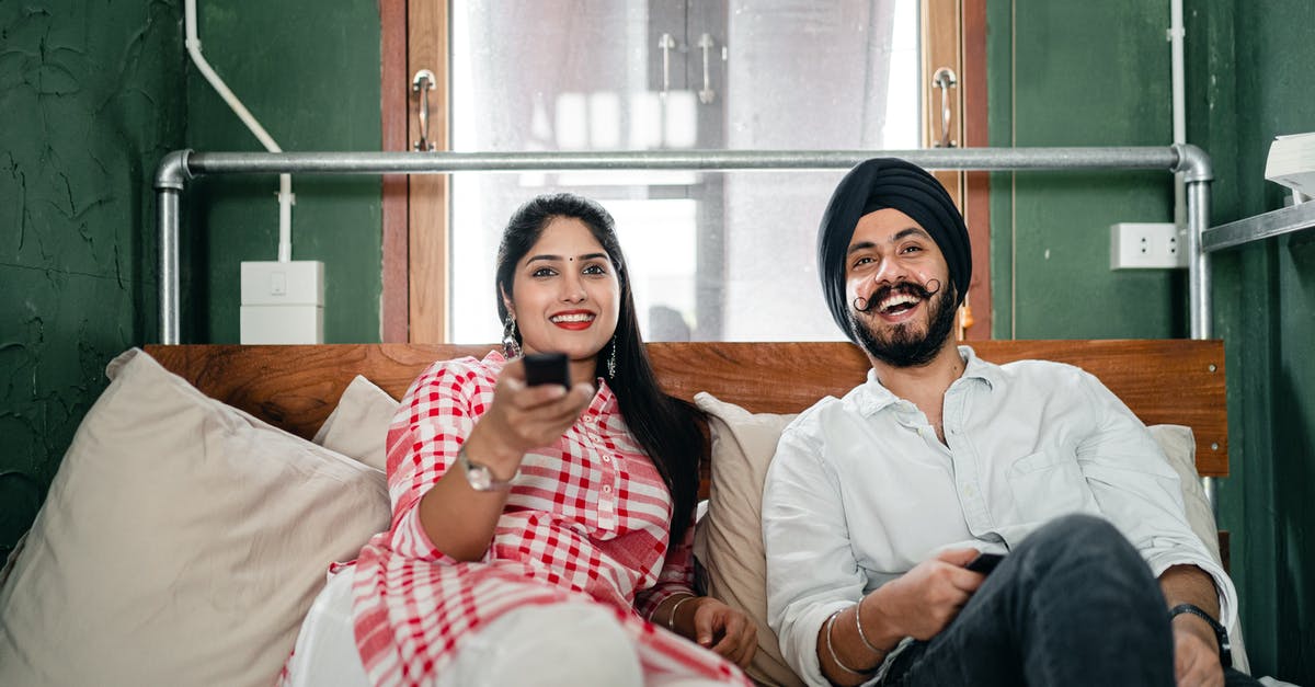 Are shows with laugh tracks considered a lower form of comedy by the TV industry? - Cheerful wife with bindi on forehead wearing plaid tunic with white trousers using TV remote control for channel switching while lying on bed with laughing Sikh husband in turban with stylish beard and twisted mustache