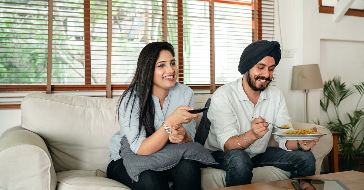 Are shows with laugh tracks considered a lower form of comedy by the TV industry? - Happy ethnic male with plate of food and female with remote control sitting on couch in living room while watching movie