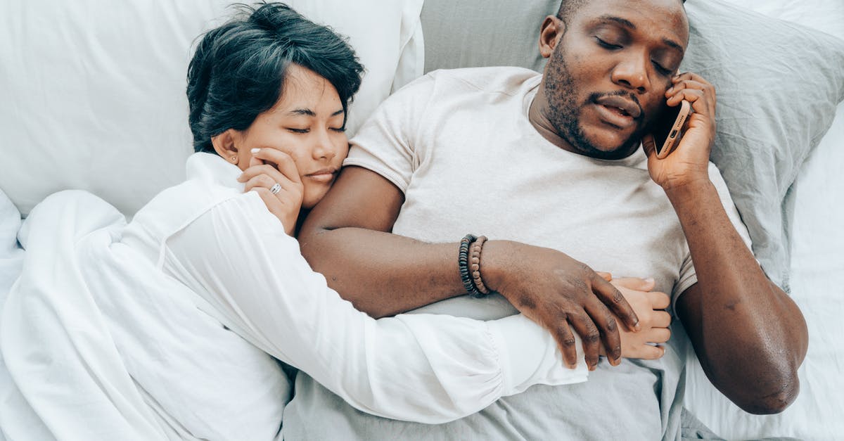Astronaut rescues corrupted wife from hell [closed] - From above of young African American man with closed eyes talking on smartphone lying in bed together with sleeping ethnic wife