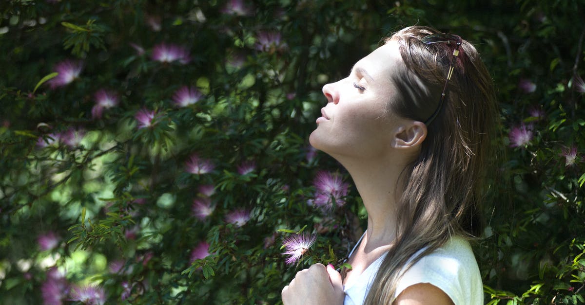 Breathing issues? - Woman Closing Her Eyes Against Sun Light Standing Near Purple Petaled Flower Plant