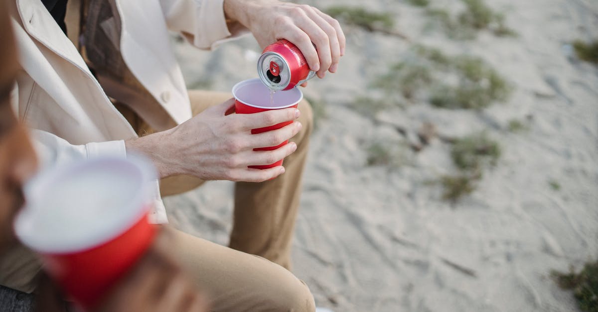 Can you really drink from Root Beer Barrels? - High angle of crop anonymous male pouring refreshing drink into red cup while sitting on sandy shore with black friend