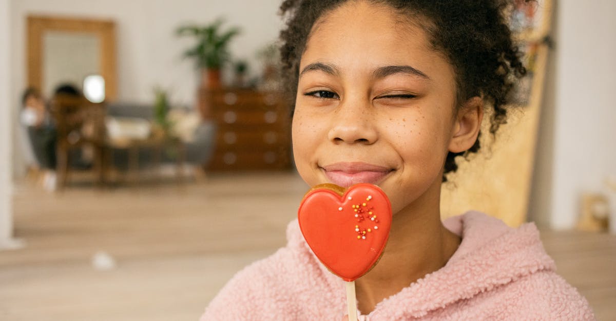 Cookie Monster's first appearance - Happy African American teenage girl with sweet heart shaped gingerbread cookie on stick winking and looking at camera in light room on blurred background