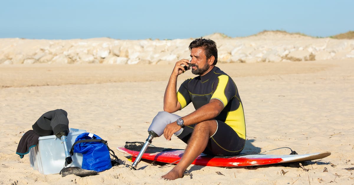 Danny Ocean using Guns - Full length tanned bearded male surfer with leg prosthesis sitting on sandy beach on surfboard and having conversation on mobile phone