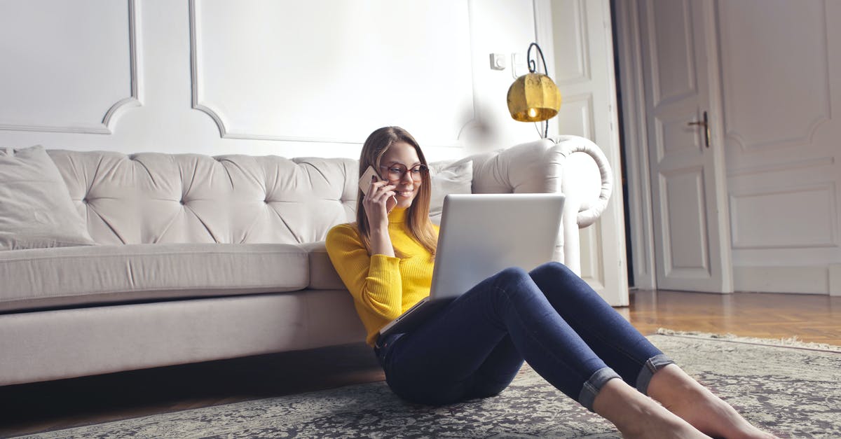 Did the McCallisters bother to call home? - Smiling barefoot female in glasses and casual clothes using laptop and having phone call while sitting on floor leaning on sofa and working on laptop against luxury interior of light living room