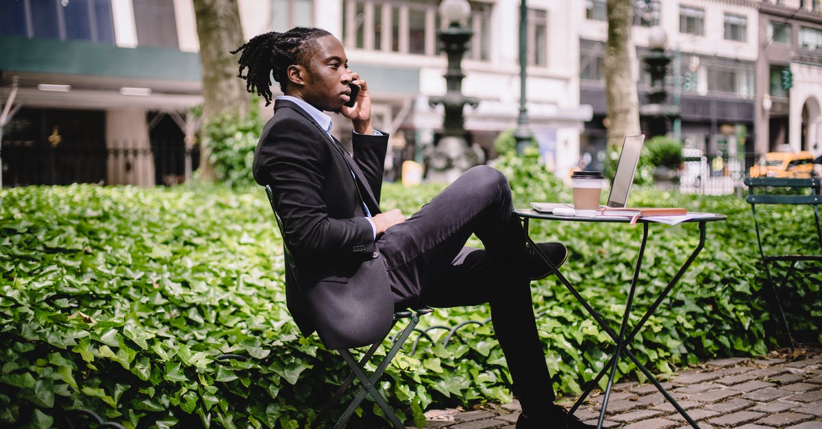 Do Saga and Martin speak Danish, Swedish or both? - Side view serious African American businessman in black formal wear sitting at table in city park with legs crossed and having conversation via modern smartphone