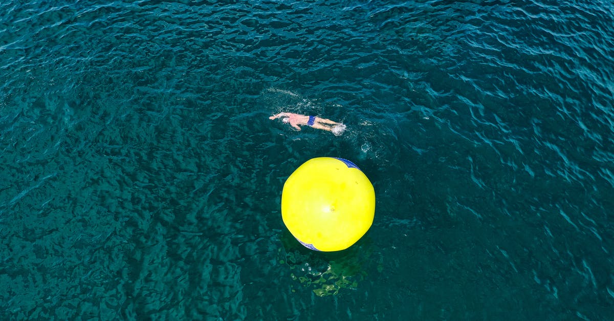 Do we ever get to know the clear motives of the aliens? - Drone view of male swimming in clear sear water around inflated yellow buoy ball