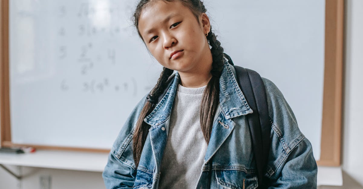Does Junior perceive he is being condescended to/not taken seriously as boss? - Concentrated Asian girl in denim jacket with backpack and hands in pockets standing in classroom against whiteboard and looking at camera
