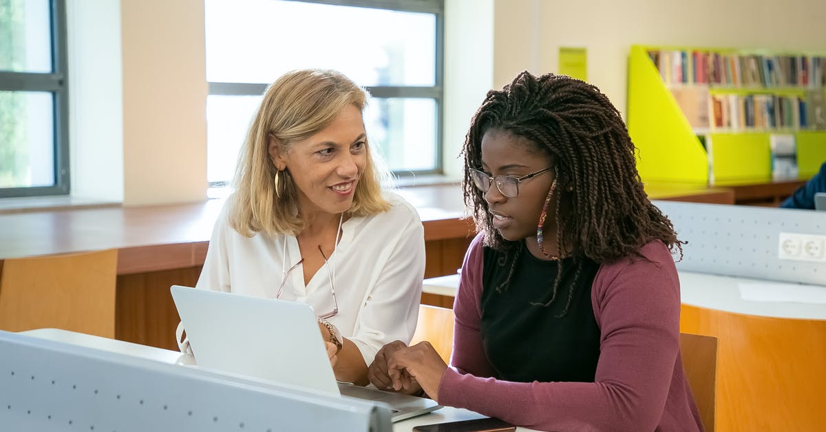 Dr. Brennan's Academic Position? - Positive adult female teacher explaining task to young black student with Afro braids doing assignment on laptop in modern classroom