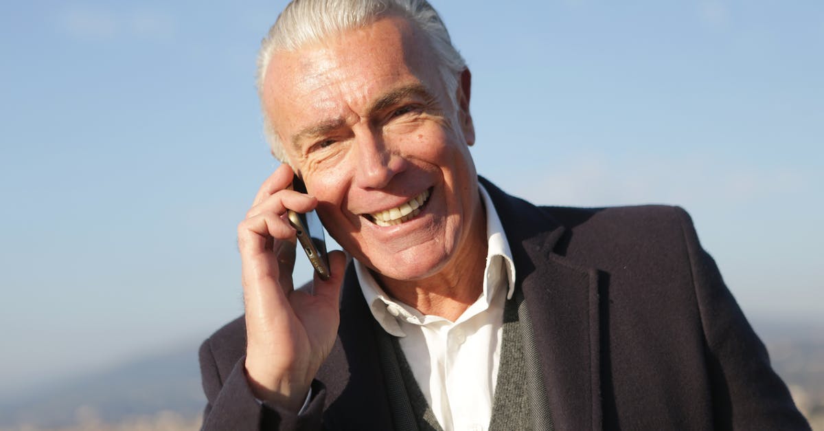 Explanation for all beings to be able to speak English? - Delighted male entrepreneur wearing classy jacket standing in city and making phone call while smiling and looking at camera
