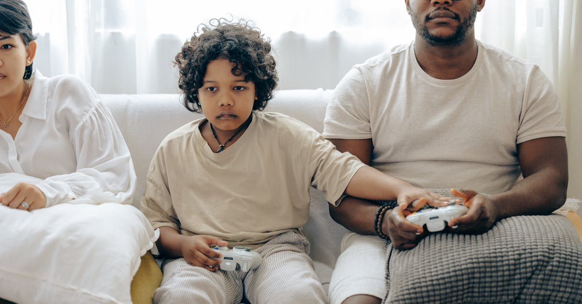 Family trapped underground/another dimension and forced to play a game show [closed] - Concentrated child showing usage of gamepad to father while playing on game console and resting on sofa together with family
