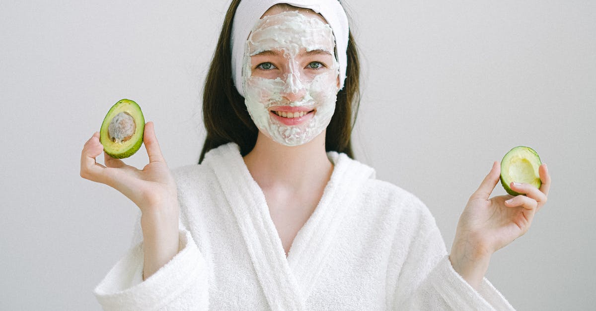 First appearance of the realistic mask effect - Cheerful female with facial mask wearing bathrobe and headband looking at camera while standing on white background with avocado in hands
