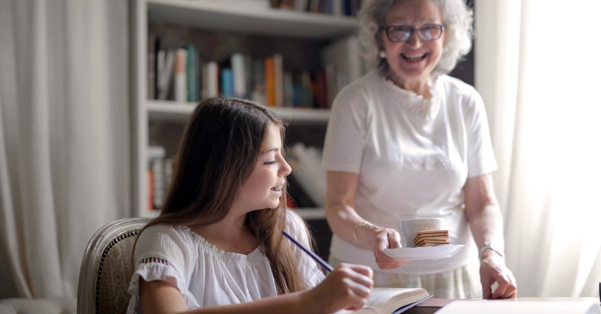 How could Edna afford to feed all those people? - Cheerful grandmother in glasses and casual clothes smiling at camera while giving pastry on plate and mug of tasty beverage to joyful teenager sitting at table with books and exercise book and study in light cozy living room