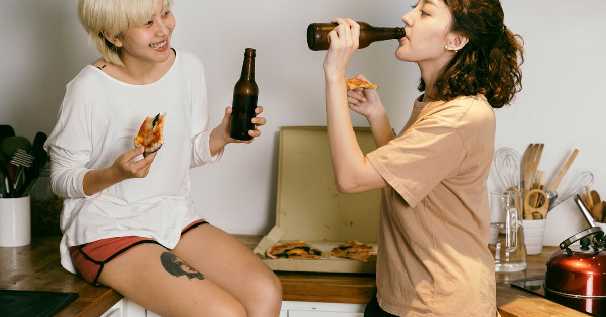 How did the other Brainiac 5 know about the bottle? - Happy tattooed blond lady eating pizza while looking at female friend in casual wear drinking beverage from bottle near kitchen utensils in apartment