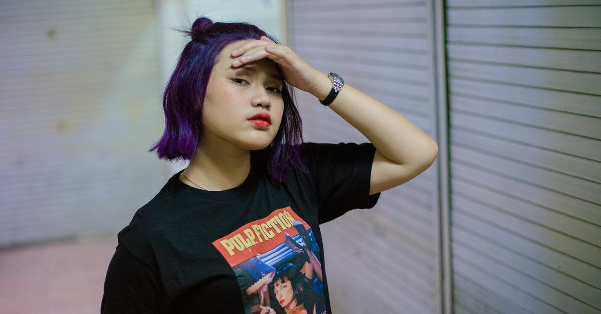 How did this character survive being bashed on the head? - Modern Asian female teenager with purple dyed hair and red lips wearing black t shirt with print keeping hand on forehead and looking at camera while standing near urban wall