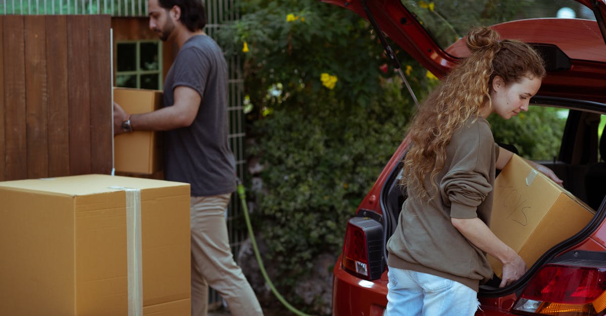 How did Tony Stark get his new suits? - Focused young woman with curly hair taking cardboard package out from red automobile trunk on street near stack of moving boxes while bearded ethnic man carrying box into new house