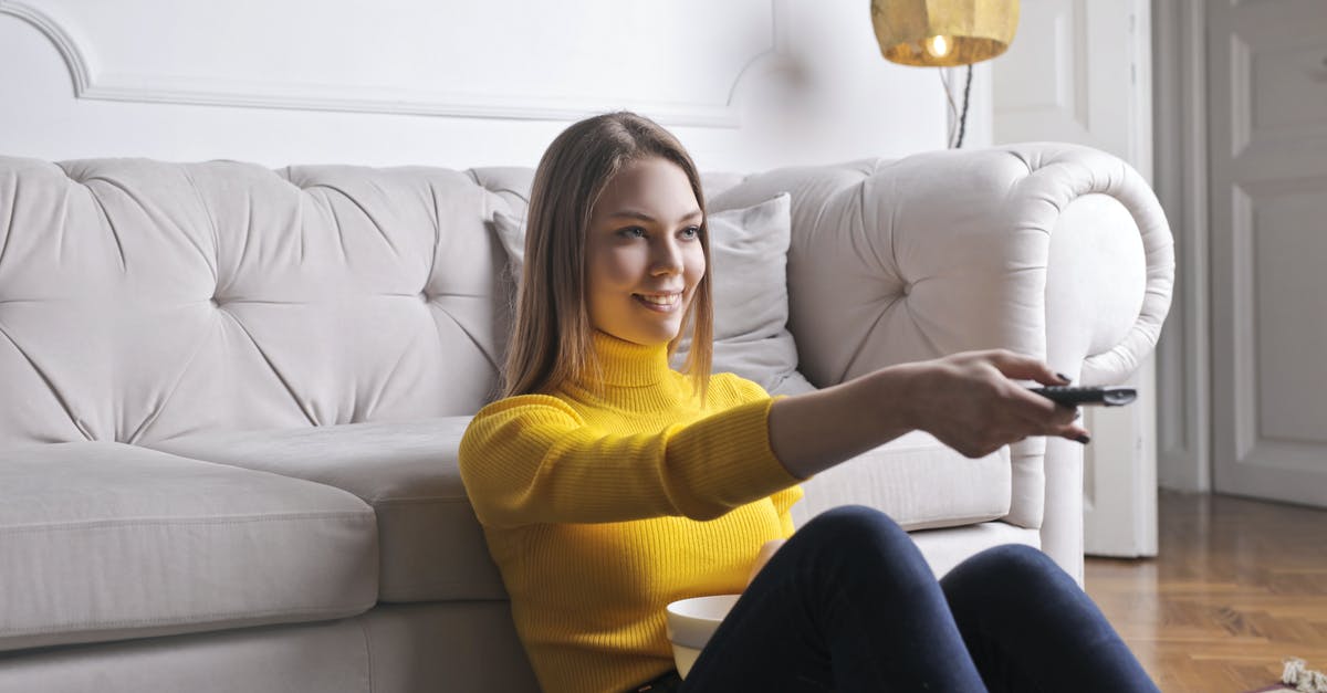 How do they film money in TV or movie? - Joyful millennial female in casual clothes with bowl of snack using remote controller while sitting on floor leaning on sofa and watching movie in cozy light living room with luxury interior