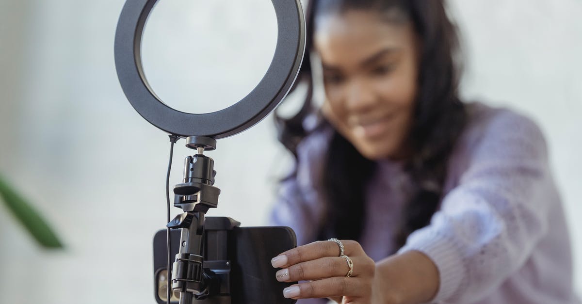 How do they shoot live commentary in reality shows? - Cheerful young African American female blogger in stylish sweater smiling while setting up camera of smartphone attached to tripod with ring light before recording vlog