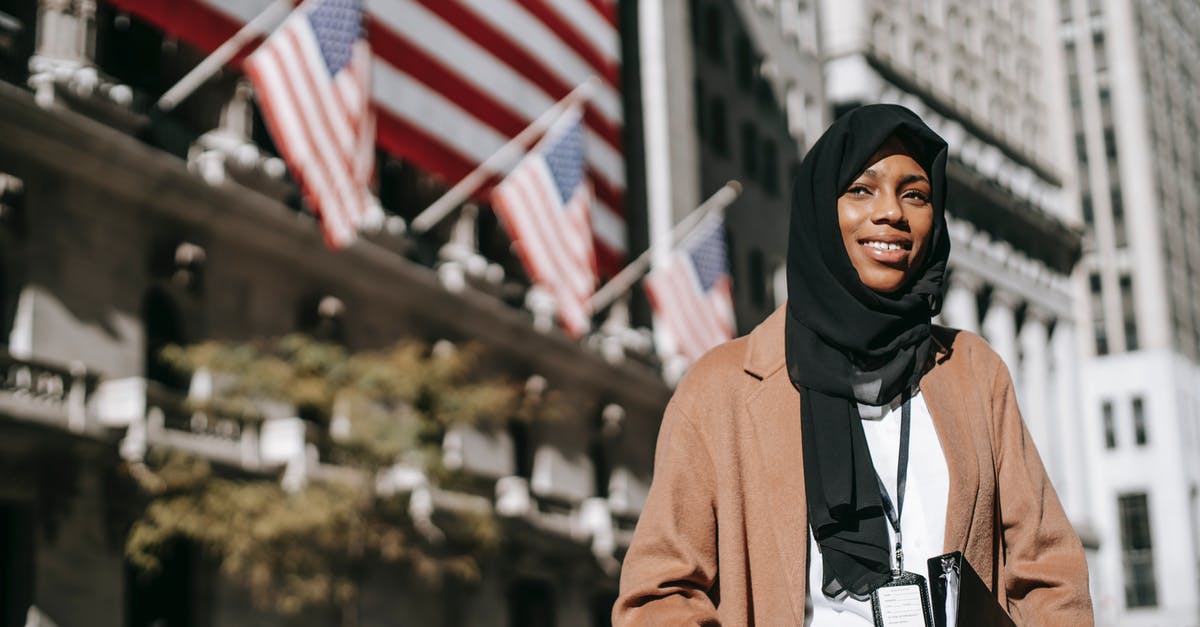 How does the veteran identify this woman as Spanish? - From below of cheerful African American female ambassador with folder wearing hijab and id card looking away while standing near building with American flags on blurred background