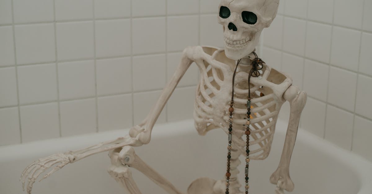 How long have they been dead? - White skeleton with long chaplet sitting in bath without water in bathroom with white tiles on wall