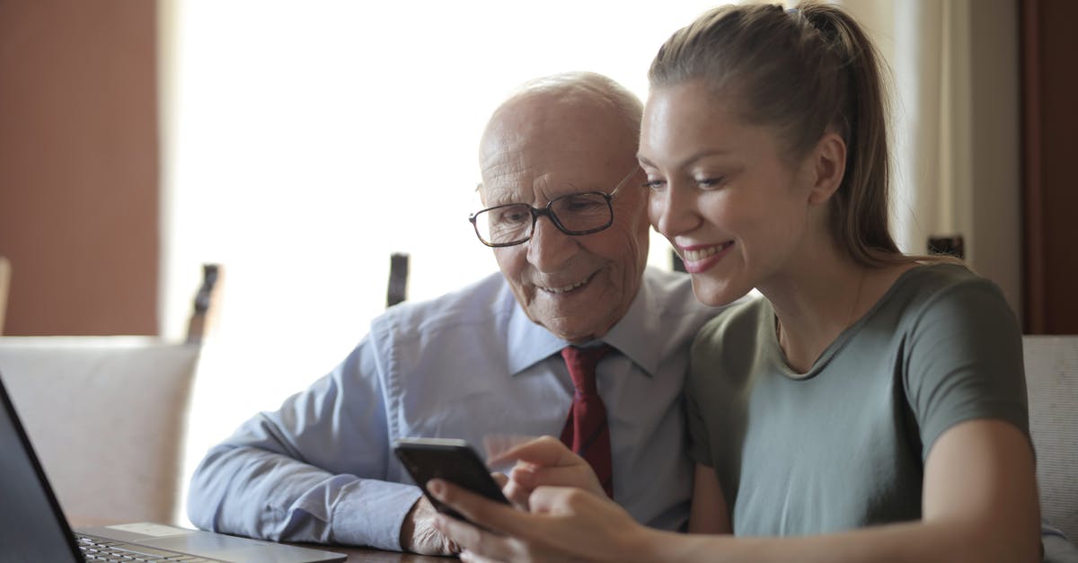 How old is Mike's granddaughter? - Smiling young woman in casual clothes showing smartphone to interested senior grandfather in formal shirt and eyeglasses while sitting at table near laptop