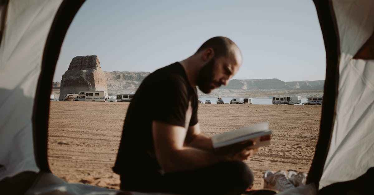 In five minutes that door is going to open, and a man... will tell me I am free to go - Side view of bearded male traveler sitting in tent with opened door and reading book in nature with parked cars in distance