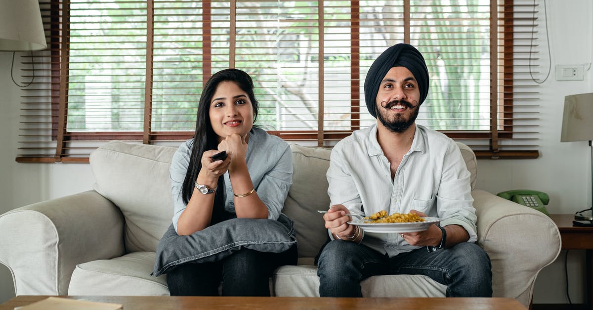 Indian movie censorship guideline - Positive male with plate of food and female in casual clothes sitting on sofa in living room and watching TV at home