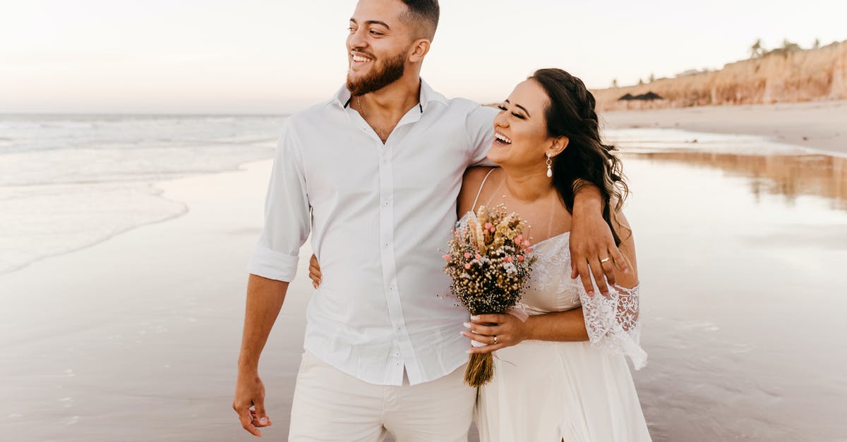 Is it common for a groom to walk the bride down the aisle? - Smiling bearded groom in white shirt and trousers walking along sandy beach together with joyful bride in wedding dress