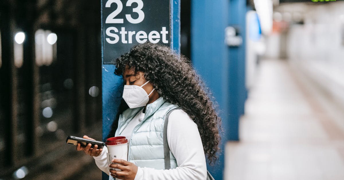 Is it normal in USA/UK to use the phone while driving? - Focused African American female wearing casual outfit and protective mask standing on New York underground platform with takeaway coffee and using mobile phone