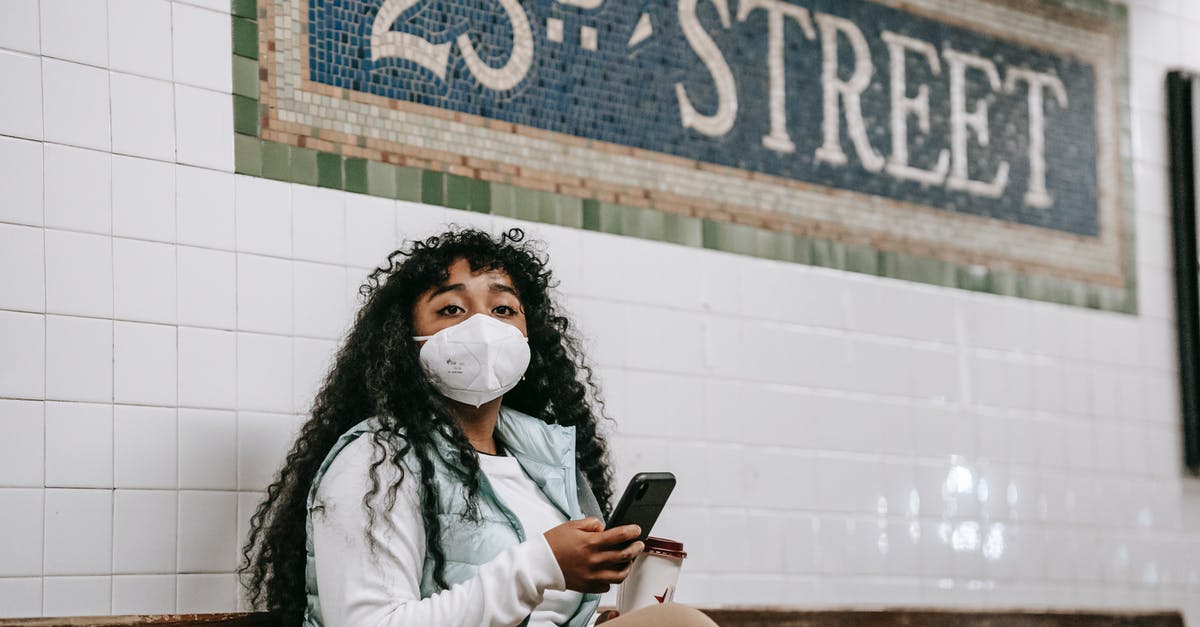 Is it normal in USA/UK to use the phone while driving? - Emotionless African American female in casual wear and protective mask using mobile phone and looking away while sitting on bench in metro station platform in New York