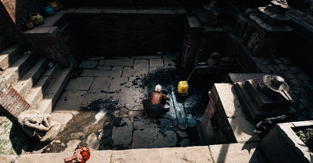 Is it realistic to build a well in the basement? - From above back view of anonymous elderly shirtless male squatting on dirty cement floor near plastic yellow container under water flow from well near stairs in old building in sunlight