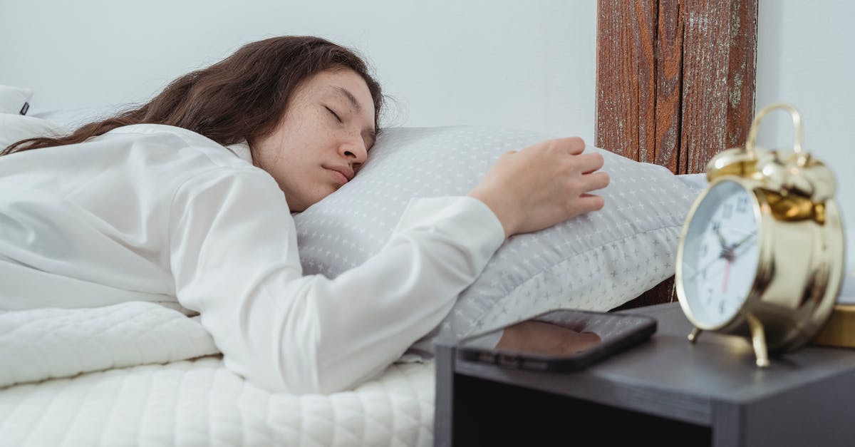 Is she lying on the phone? - Young woman with dark long wavy hair sleeping peacefully on belly on comfortable bed under white blanket near bedside table with alarm clock and smartphone