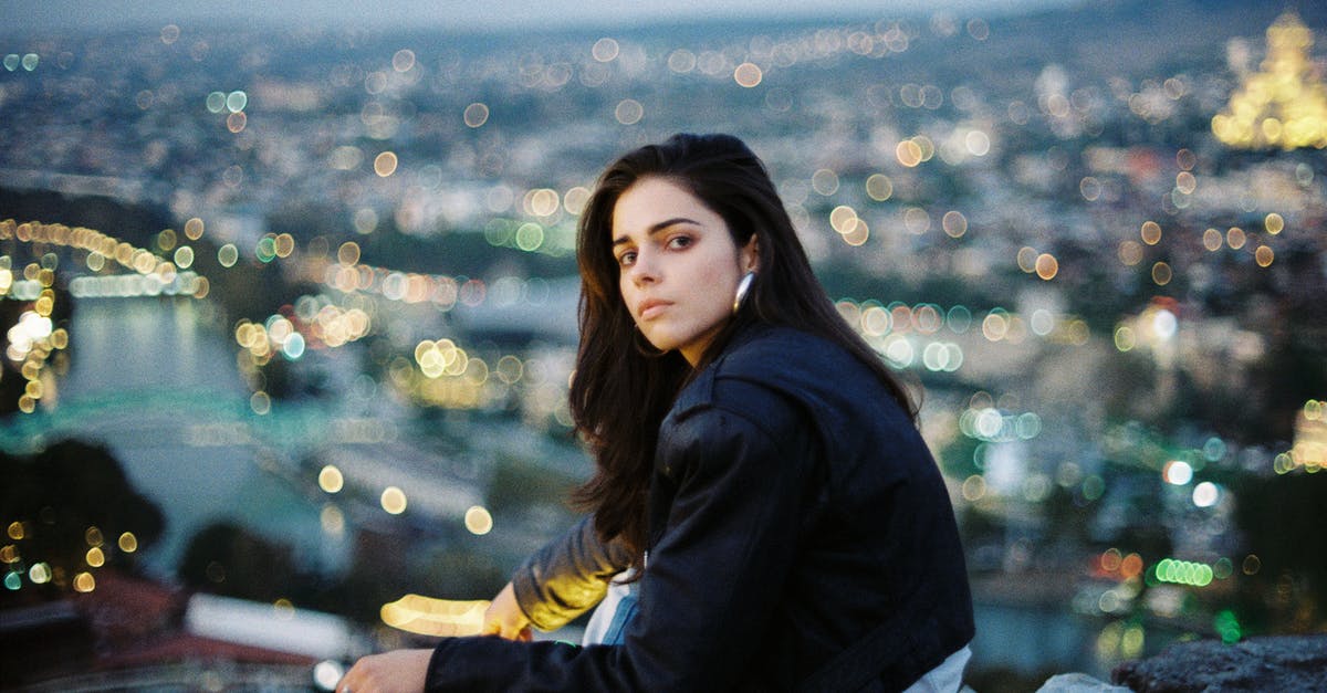 Opening scene with a woman who gets her high heels stuck in a grate [closed] - Emotionless young female in black jacket and jeans sitting on rooftop above city street in evening time and looking at camera