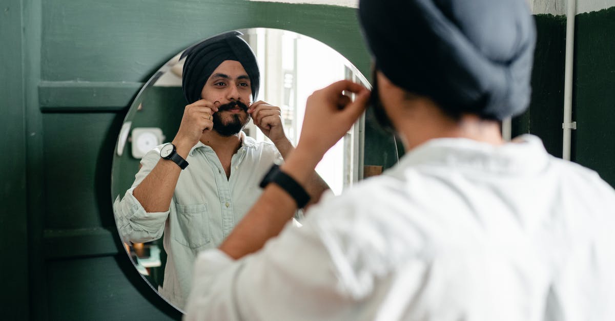 Origin of the Mustache Twirling Villain - Back view of handsome young Indian male hipster with twisted mustache wearing denim shirt and traditional turban standing against mirror on wall while taking care about appearance