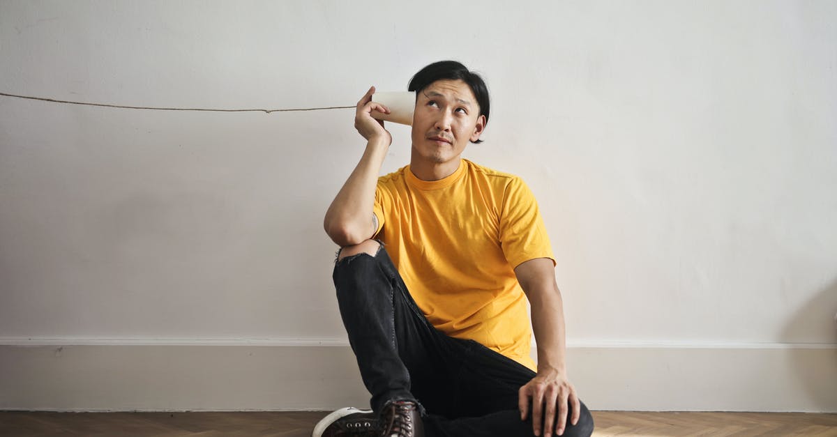 Phone calls to the past - The Long Game - Asian guy in casual clothes using paper cup with thread as telephone while sitting on floor against white wall