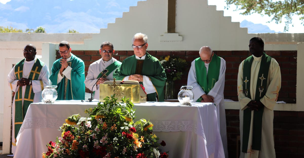 Priest avoids turning into a vampire by exposing himself to sunlight [closed] - Elderly priest in eyeglasses and catholic vestment standing with prayer hands near table with Bible and multiethnic deacons during liturgy process behind mountains in sunlight