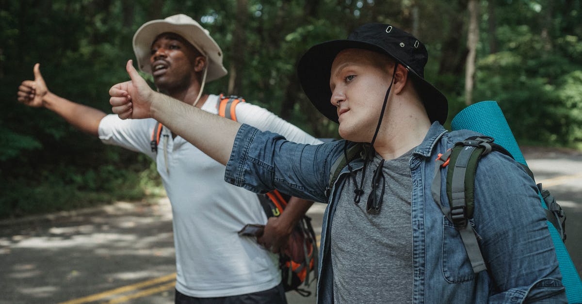 "I like your outfit. Affordable?" How is this an insult? - Young multiracial male backpackers in panama hats with hiking equipment standing with thumbs up on roadway against forest and looking away