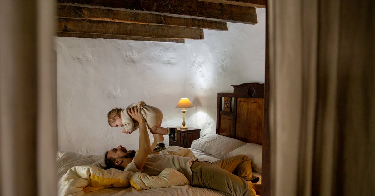 "No Country for Old Men", why is it called so? - Young father wearing casual clothes lying on back on bed and carrying adorable little child on rising hands in bedroom with rough white walls and ceiling beams while spending time at home together