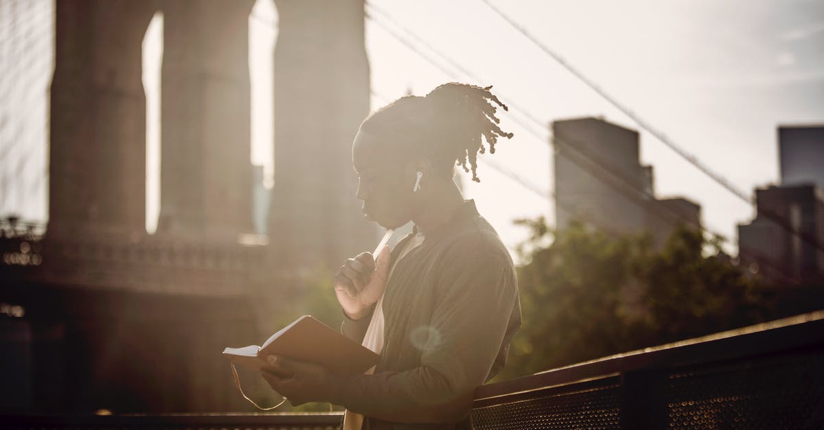 Songs overlap in the Jungle Book - Side view of pensive young black man with braided hairstyle reading book and listening to music with wireless earphones while standing on city promenade against bridge