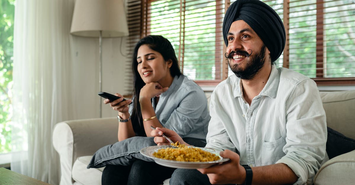 TV movie where an inconspicous truck transporting valuables is intercepted by terrorists in a dead radio spot [closed] - Positive young bearded man in casual wear and turban eating traditional saffron rice on sofa while wife switching channels on TV with remote controller