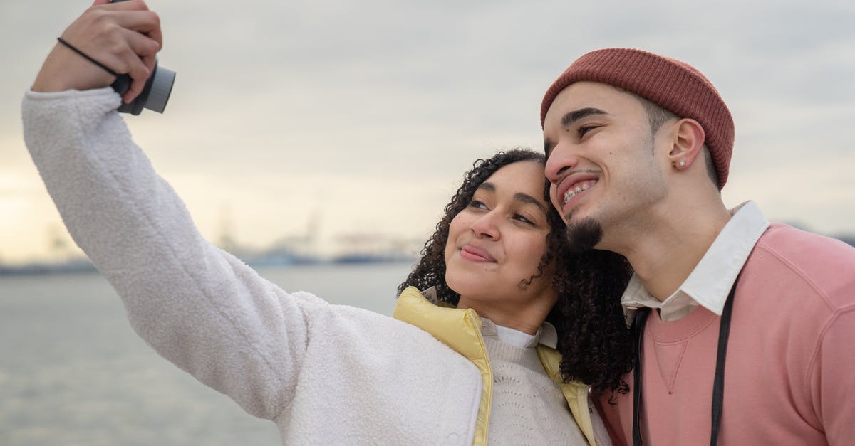 Use of anachronisms in film intros to surprise the user with the revelation of the actual time period - Smiling Latin American couple in warm clothes standing near river while taking self photo on analog camera in daylight under cloudy gray sky