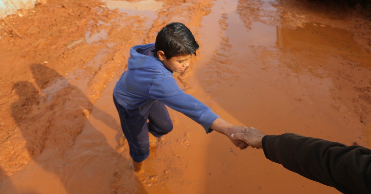 What did the aliens need help for? - High angle of crop person holding hands with ethnic boy stuck in dirty puddle in poor village