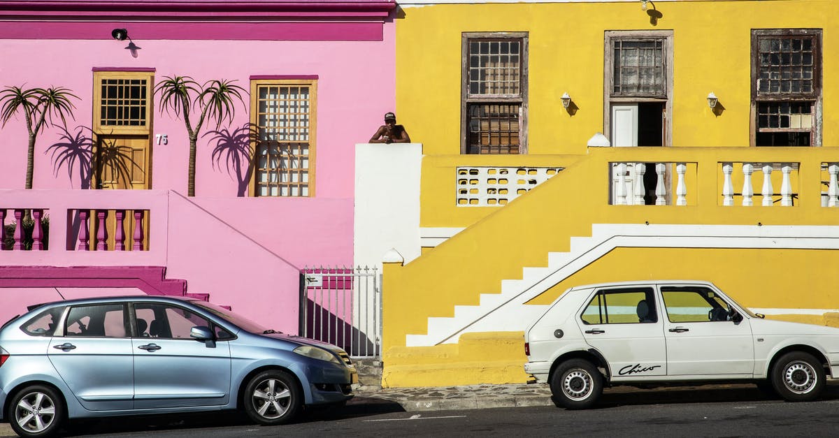 What is the main trait between each major house? - Distant African American man standing between pink and yellow residential buildings located near road with parked cars on street in town