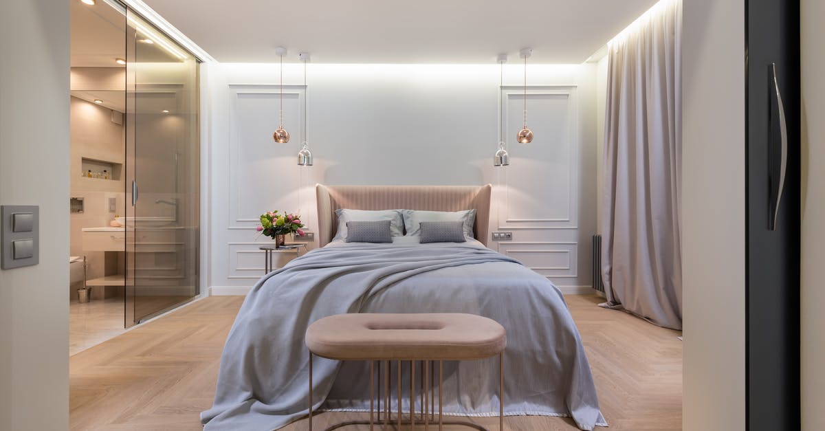 What is the object found by the bed? - Interior of modern light home with bed with pillows and blanket next to table under lamps near pouf and glass door to washroom