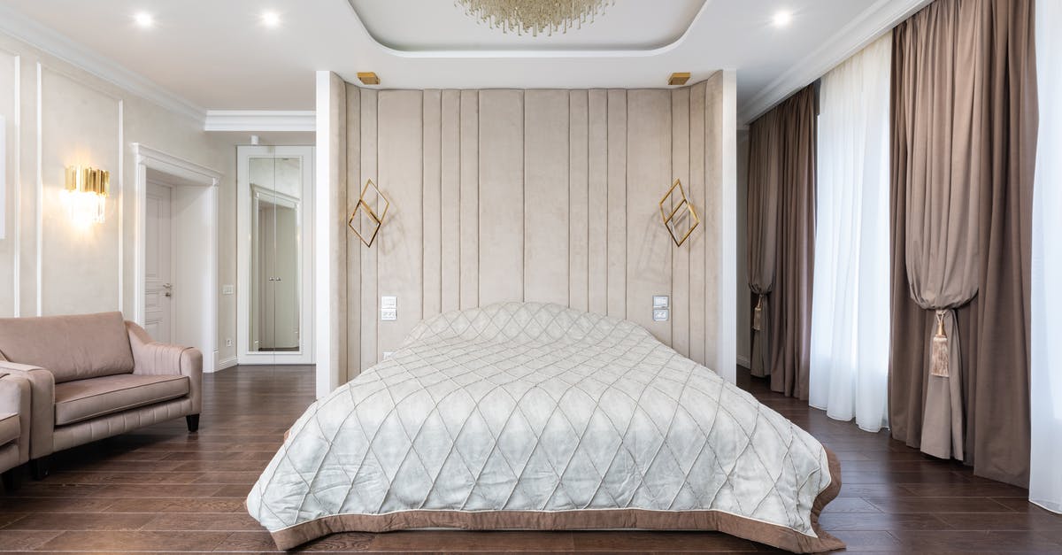 What is the object found by the bed? - Interior of modern bedroom with bed next to armchairs under chandelier with curtains on windows