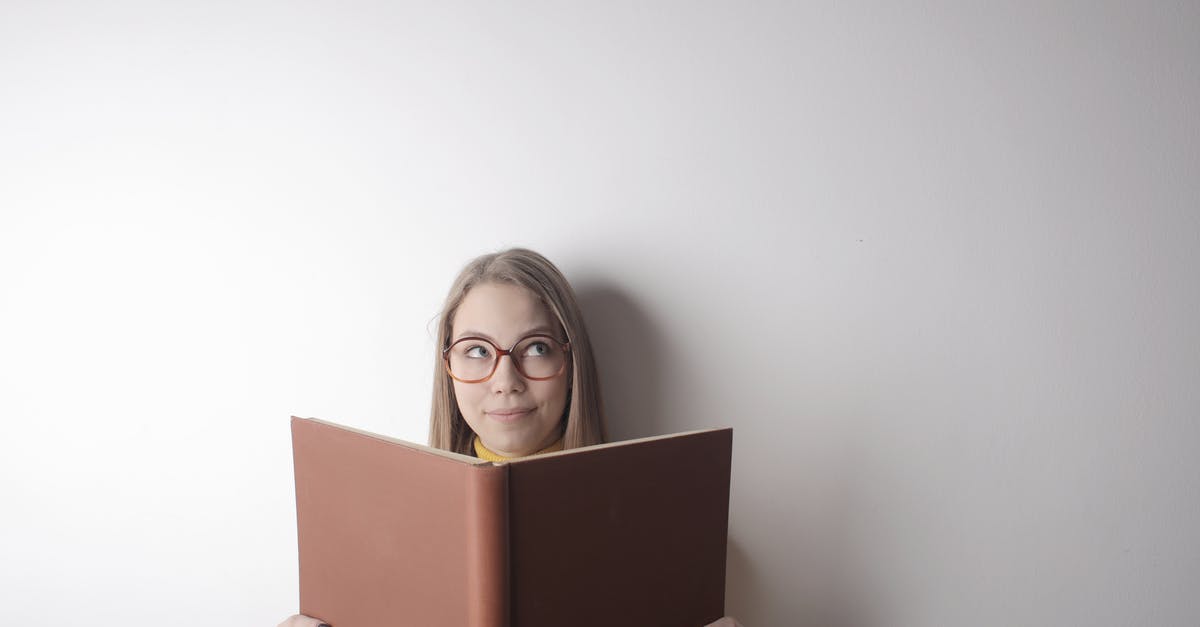 What was up with the lady in library? - Positive young curious female wearing eyeglasses looking up while standing against gray wall with big opened book with brown cover in hands