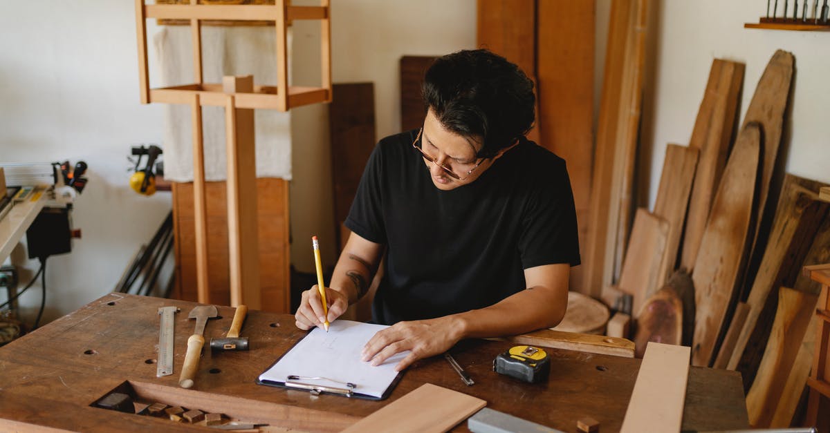 Who's in on the master plan? - Concentrated man in glasses sitting at wooden table in workshop and holding pencil while creating new object on paper